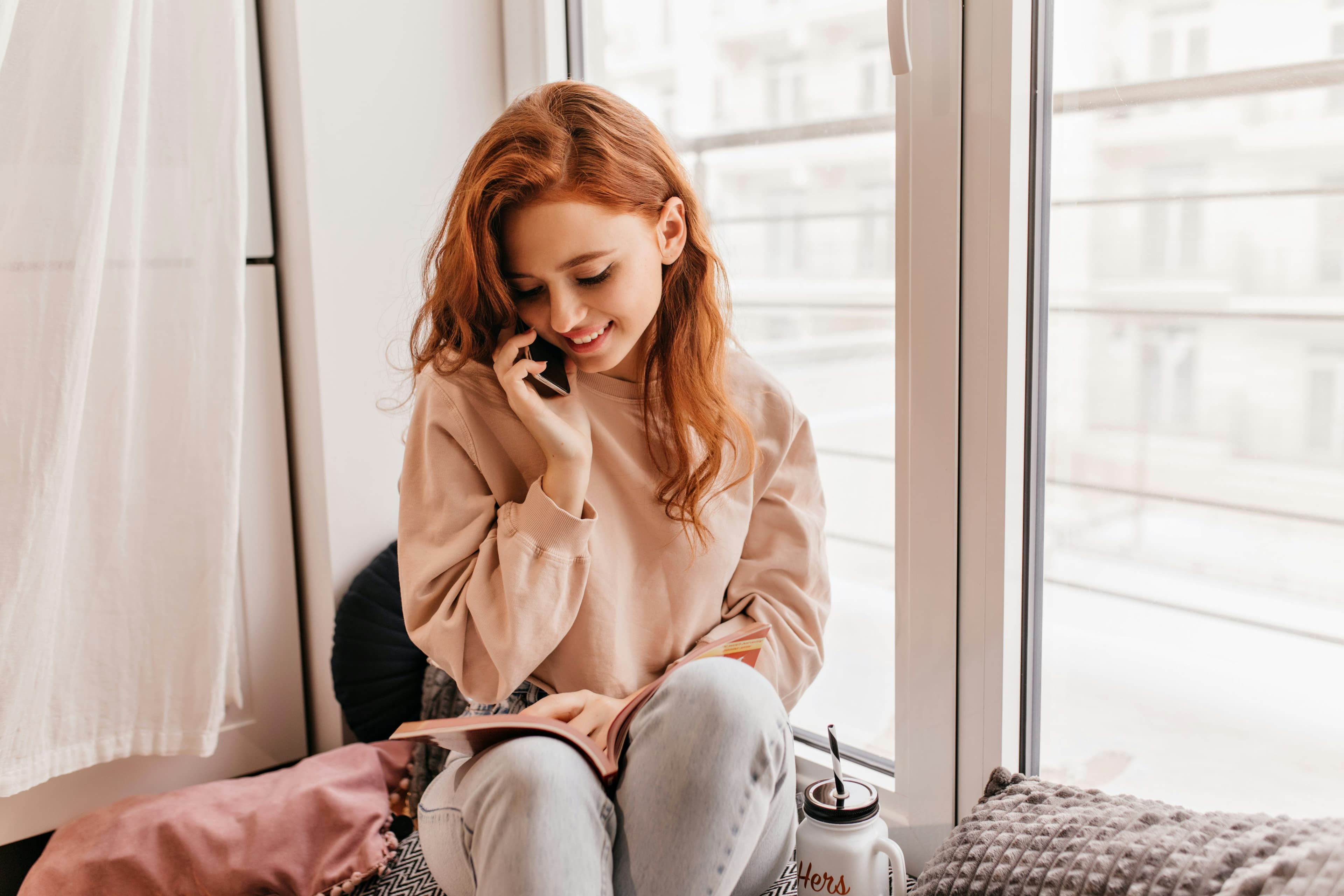 A young woman seated on a window bench in bright daylight, smiling softly mid-conversation with a phone to her ear.